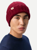 A close-up of a man wearing a dark red ribbed knit beanie with a small beige brand patch on the front, paired with a navy blue crewneck sweater against a light gray background. #color_bordeaux