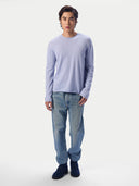 A man modeling a light purple long-sleeve crewneck sweater, paired with light blue jeans and dark blue suede boots, against a solid light gray background.
