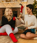 Two women exchanging cashmere Christmas gifts by a fireplace, wearing black and cream jumpers with red socks