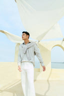 Model in grey shirt and white trousers with knit draped over shoulders, a cashmere travel outfit by the sea