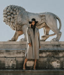 Model in long open beige coat and black hat showing autumn cashmere essentials, front view outdoors by stone lion