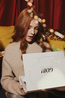 Woman in beige ribbed cashmere jumper opening a white GOBI gift box, festive lights behind, cashmere gift ideas