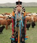 Woman in traditional Mongolian costume and headdress standing among goats on green steppe, things to do in Mongolia