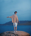Model on a lakeside rock in a beige textured cashmere poncho jumper and white skirt, showing the timeless appeal of cashmere