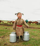 Smiling Mongolian child in a traditional deel carrying two milk churns on the steppe, with horses behind