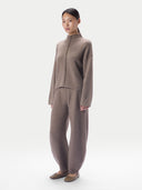 A model wearing a matching taupe knit two-piece set consisting of a mock-neck long-sleeve top with a braided front detail and wide-leg trousers, paired with brown suede flats, against a light gray studio background.#color_taupe