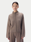 A model wearing a taupe brown knit sweater with a braided front detail and matching knit pants, photographed against a clean light gray studio background.