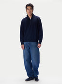 A man wearing a navy blue quarter-zip pullover sweater and loose-fit blue jeans, standing against a plain light gray studio background.