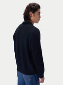 A man shown from the back wearing a black knit sweater and blue jeans, photographed against a clean light gray studio background.