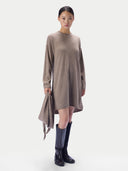 A model wearing a taupe-brown long-sleeve knit dress with an asymmetrical draped hem, paired with dark knee-high boots, against a plain light gray studio background.