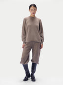 A model stands against a light gray studio background wearing a taupe knit sweater and matching wide-leg cropped pants, paired with dark knee-high boots.