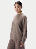 A model wearing a taupe knit sweater with textured cable detailing on the sleeves, paired with matching knit pants, against a light gray studio background.