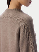 Close-up of a person wearing a taupe knit sweater, showing the back and shoulder with a braided cable pattern and ribbed crew neck.