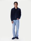A man modeling a dark navy blue zip-up jacket, light-wash blue jeans, and navy blue slippers against a plain white studio background.