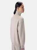A woman shown in side profile wearing a light beige high-neck knit sweater against a plain light gray studio background.
