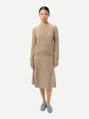 A model stands against a light gray studio background wearing a matching beige knit sweater and midi skirt set, paired with gray slip-on shoes.