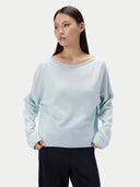 A woman modeling a light mint green knit sweater with a wide boat neckline and long sleeves, paired with dark charcoal pants, against a plain light gray studio background.