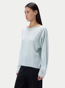 A woman modeling a light mint green knit sweater with long sleeves and a wide boat neckline, paired with black trousers, against a plain light gray studio background.