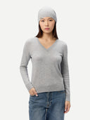 A woman wearing a light gray long-sleeve V-neck sweater, a matching gray knit beanie, and blue jeans, standing against a plain light gray studio background. #color_light-gray