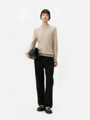 Person wearing a beige sweater and black pants holding a black clutch against a white background. #color_soft-beige