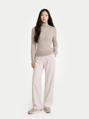 Woman wearing a beige turtleneck sweater and white pants on a white background