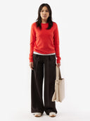 Woman wearing a red sweater and black pants holding a beige bag on a white background. #color_red-orange