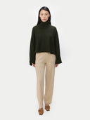 A model stands against a light gray studio background wearing a dark olive green cropped turtleneck sweater, beige wide-leg trousers, and brown shoes.