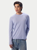 A man wearing a light lavender long-sleeve crewneck knit sweater and blue jeans, standing against a plain light gray studio background.