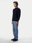 A man wearing a dark navy blue mock neck sweater, light blue jeans, and black leather shoes, standing in profile against a light gray studio background.