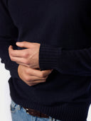 A close-up of a person wearing a dark navy blue knit sweater, showing the ribbed cuffs and hands, paired with blue jeans and a brown belt.