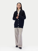 A model stands against a light gray studio background wearing a dark navy cardigan over a cream collared shirt, paired with light beige wide-leg trousers and dark shoes.