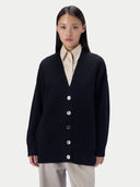 A model wearing a black knit cardigan with silver buttons over a cream collared shirt and light beige trousers, shot against a solid light gray background.