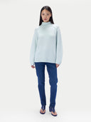 A model stands against a light gray studio background wearing a pale icy blue mock-neck knit sweater, dark blue jeans, and dark blue heeled sandals.