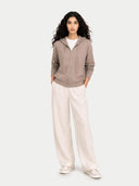 Woman wearing a brown zip-up hoodie and white pants on a white background.