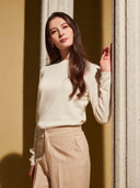 Woman in a light-colored sweater and beige pants standing against a wall with sunlight casting shadows. #color_white
