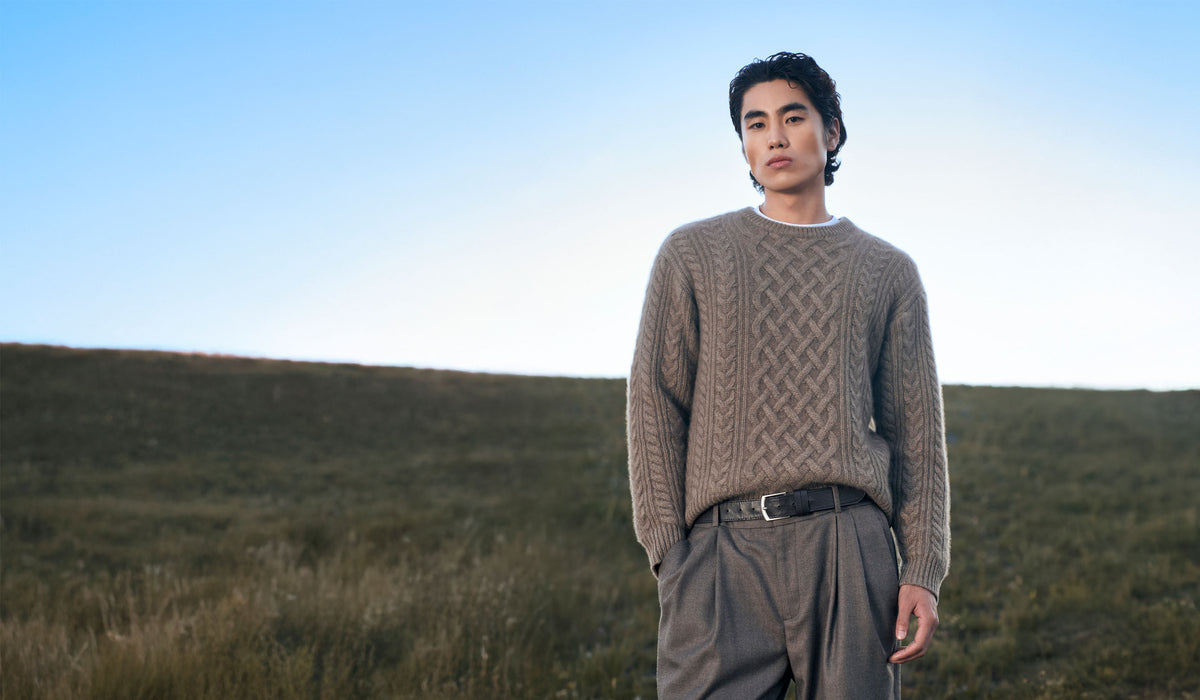 Man in a taupe cable-knit cashmere jumper and grey trousers standing on a grassy hillside under a blue sky
