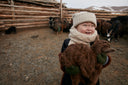A laughing Mongolian child in a knitted hat hugging a baby goat inside a herd pen