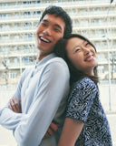 A laughing couple standing back to back in the city, in a pale blue cashmere polo and a navy patterned cashmere dress