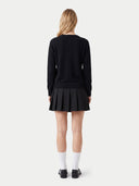 A model shown from the back wearing a black long-sleeve sweater and a black pleated mini skirt, paired with white knee-high socks and black shoes, against a light gray studio background.
