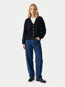 A woman modeling a black button-front cardigan over a white top, paired with blue jeans and black shoes, against a plain light gray studio background.