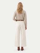 A woman shown from the back wearing a beige cable-knit sweater, off-white wide-leg trousers, a thin brown belt, and brown ankle boots against a plain light gray studio background.