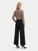 A woman is shown from the back wearing a cropped taupe-brown turtleneck sweater, black wide-leg trousers, and black high heels against a light gray studio background.