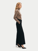 A woman stands in profile wearing a taupe-brown ribbed turtleneck sweater, a high-waisted black wide-leg skirt, and black pointed-toe heels against a light gray studio background.