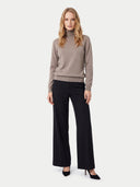 A woman modeling a taupe turtleneck sweater and black wide-leg trousers against a light gray studio background.
