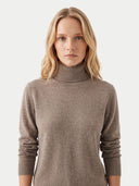 A woman with blonde hair wearing a taupe brown knit turtleneck sweater, photographed against a clean off-white background.