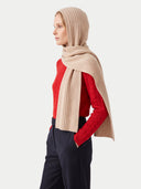 A woman in profile wearing a beige ribbed hooded hat draped over a red knit sweater and dark navy trousers, photographed against a light gray studio background.
