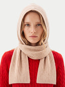 A woman wearing a ribbed knit hooded hat n a light beige color, styled over a bright red cable-knit sweater against a light gray background.