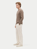 side view of a man standing wearing a taupe color crew neckline ribbed sweater and white pants on a light gray background.