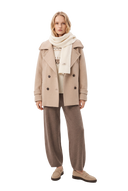 Woman in a taupe trench-style cashmere peacoat over a white Nordic jacquard jumper, with a soft white wide rib knit scarf, taupe barrel-leg knit trousers and beige loafers