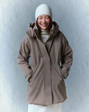 A woman in a taupe CashmereTech parka and white beanie against a white wall